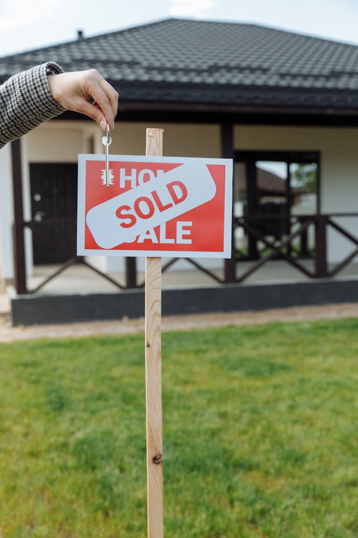 gallery-1 A hand holding a key in front of a sold sign and a house, symbolizing a successful real estate transaction.